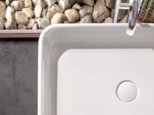 Rechteckiges Stahl-Email-Waschbecken auf Einbauplatte, weiße Oberfläche, moderner Wasserhahn, Kieselsteine im Hintergrund.
